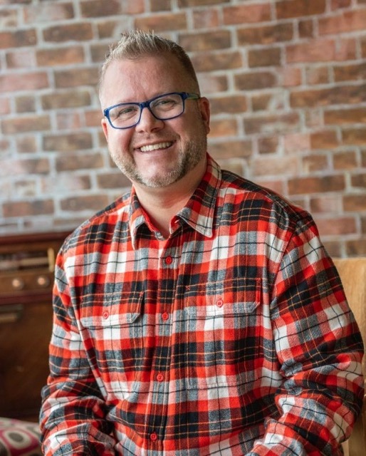 Headshot of Michael, a smiling bearded man with glasses wearing a red and black checkered shirt in a casual indoor setting.