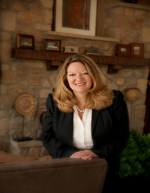 Headshot of Michelle Claymore, a smiling woman with long wavy light brown hair, wearing a black blazer, white blouse, and pearl necklace, seated in a cozy living room with a stone wall, framed photos, and decorative accents in the background.
