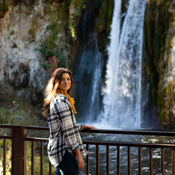 Girl next to railing looking back at camera in front of waterfall.