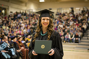 BHSU graduate holding their diploma and smiling, audience and other graduates in the background
