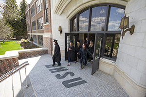 BHSU Graduates walking out the front doors of Woodburn Hall in their cap and gowns