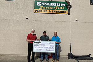 John Heck, Clair Donovan, Dr. David Berberick holding a giant check outside of the Stadium Sports Grill