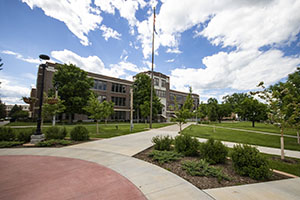 Picture of the BHSU campus green, lots of green grass and trees, Woodburn Hall can be seen in the background with a blue and cloudy sky