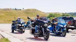 A group of motorcyclists on their bikes in a parking lot with rolling hills in the background; the lead rider gives a peace sign to the camera.