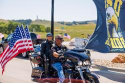 A man and a woman on a motorcycle with three American flags attached to the back, smiling as they participate in the BHSU Jacket Ride.