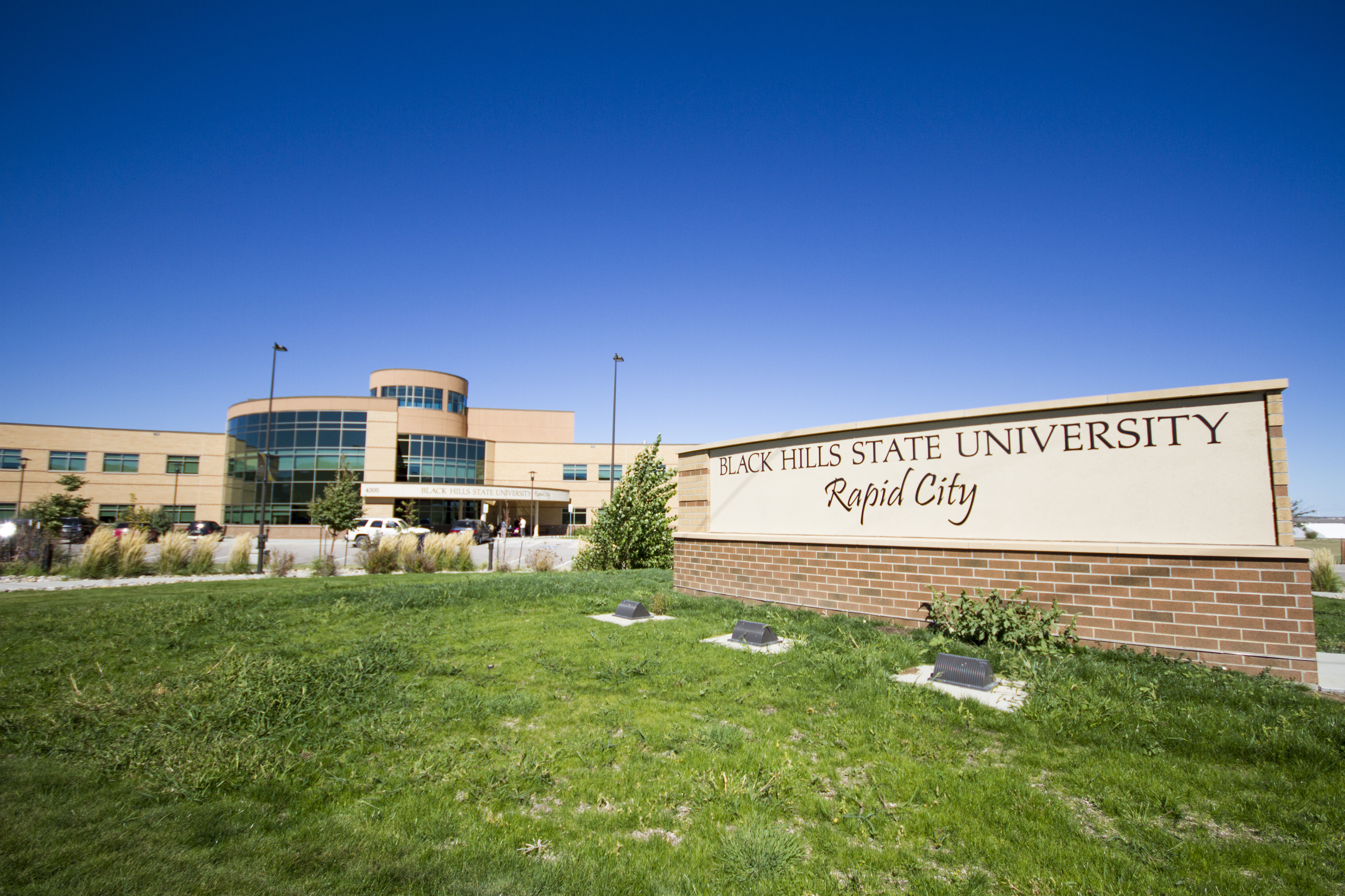 Exterior view of Black Hills State University-Rapid City campus featuring a modern building with large windows and a curved facade, set against a clear blue sky. In the foreground, a low brick wall displays the university's name in prominent lettering.