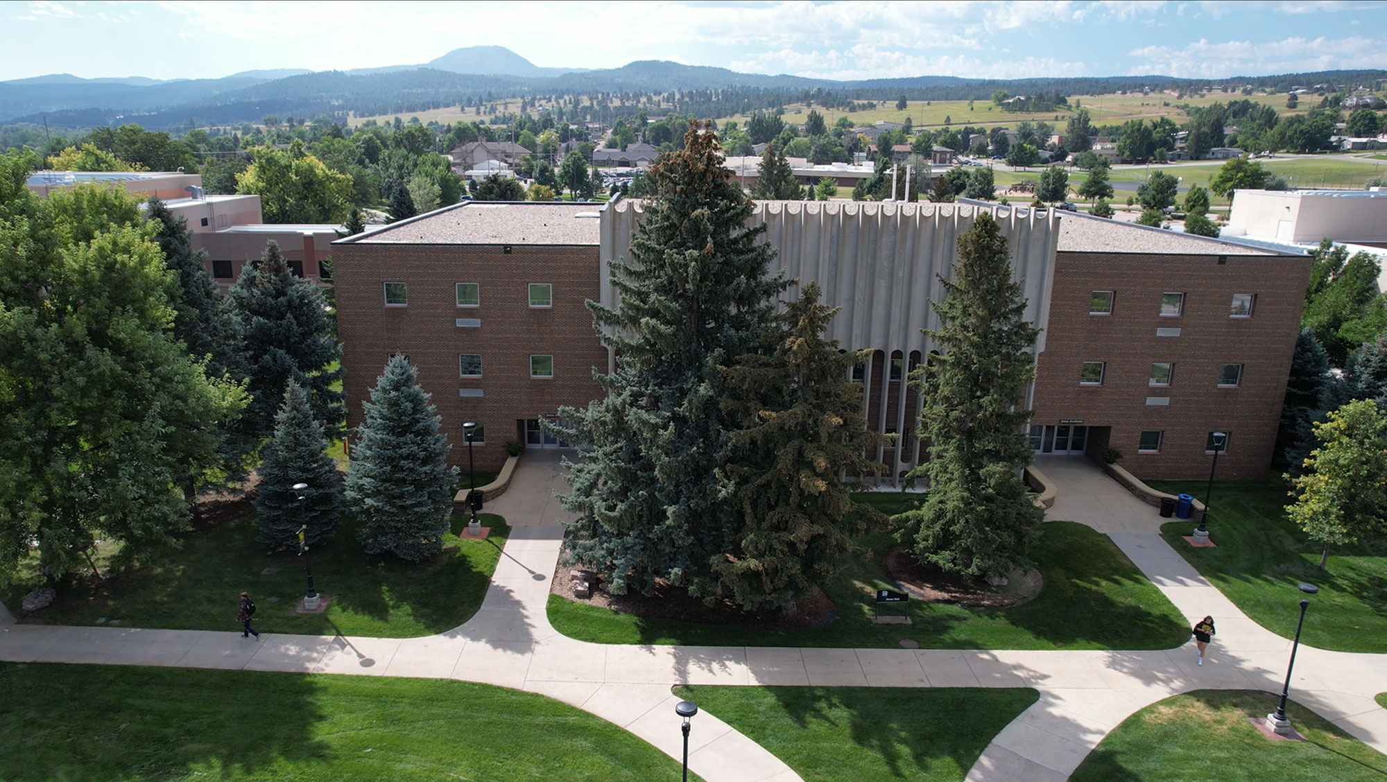 Aerial view of a Jonas Academic Hall on the BHSU campus with brick buildings surrounded by lush green trees, with mountains visible in the distance. People are walking along the pathways.