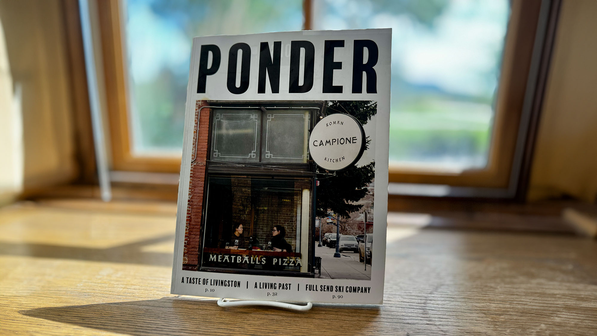 A magazine titled "PONDER" is displayed on a table, featuring images of urban life including an image of restaurant. The scene is backlit by a sunlit window.