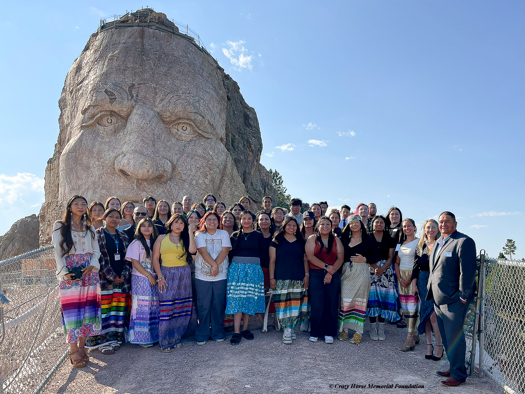 The 2024 cohort of students and staff of the Black Hills State University and The Indian University of North America® (IUNA) at Crazy Horse Memorial® 7TH GEN.® summer program pose for a picture in front of the iconic monument. 