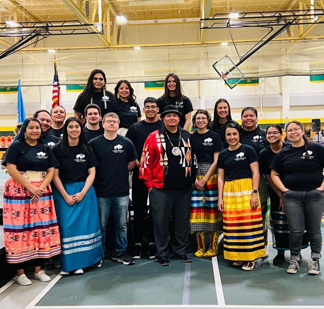 A group of people in matching black shirts, some in colorful ribbon skirts, pose in a gym. A man in the center wears a red patterned jacket and medallion.