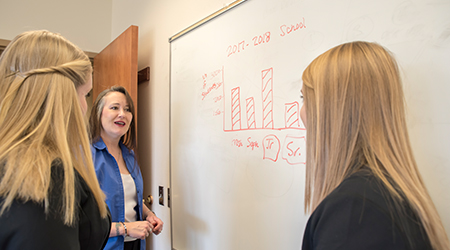 Three people, two women and a man, are gathered in an office, looking at a whiteboard. The man in the middle, wearing a blue shirt, is pointing at a bar graph drawn on the board. The two women are facing the board and the man, engaged in a discussion. The whiteboard has a bar graph with 'Freshman,' 'Sophomore,' 'Jr,' and 'Sr' written underneath the bars.