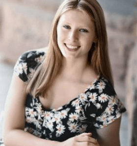 A young woman with long blonde hair sits on a bench outdoors. She is wearing a black top with white daisies and is smiling at the camera with her hands clasped in her lap.