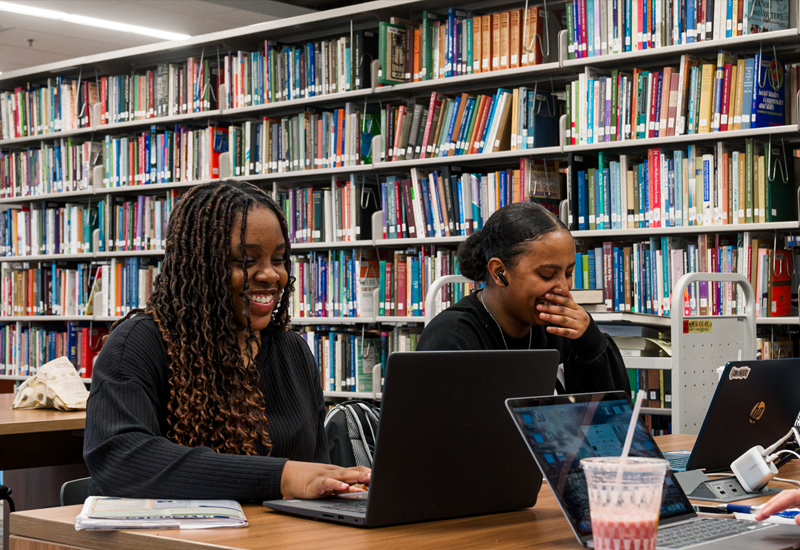 Two students study in the library with bookshelves in background.