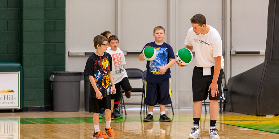 PE instructor demonstrating ball skills to elementary school children in a gymnasium, showcasing the hands-on teaching approach of the Physical Education program.