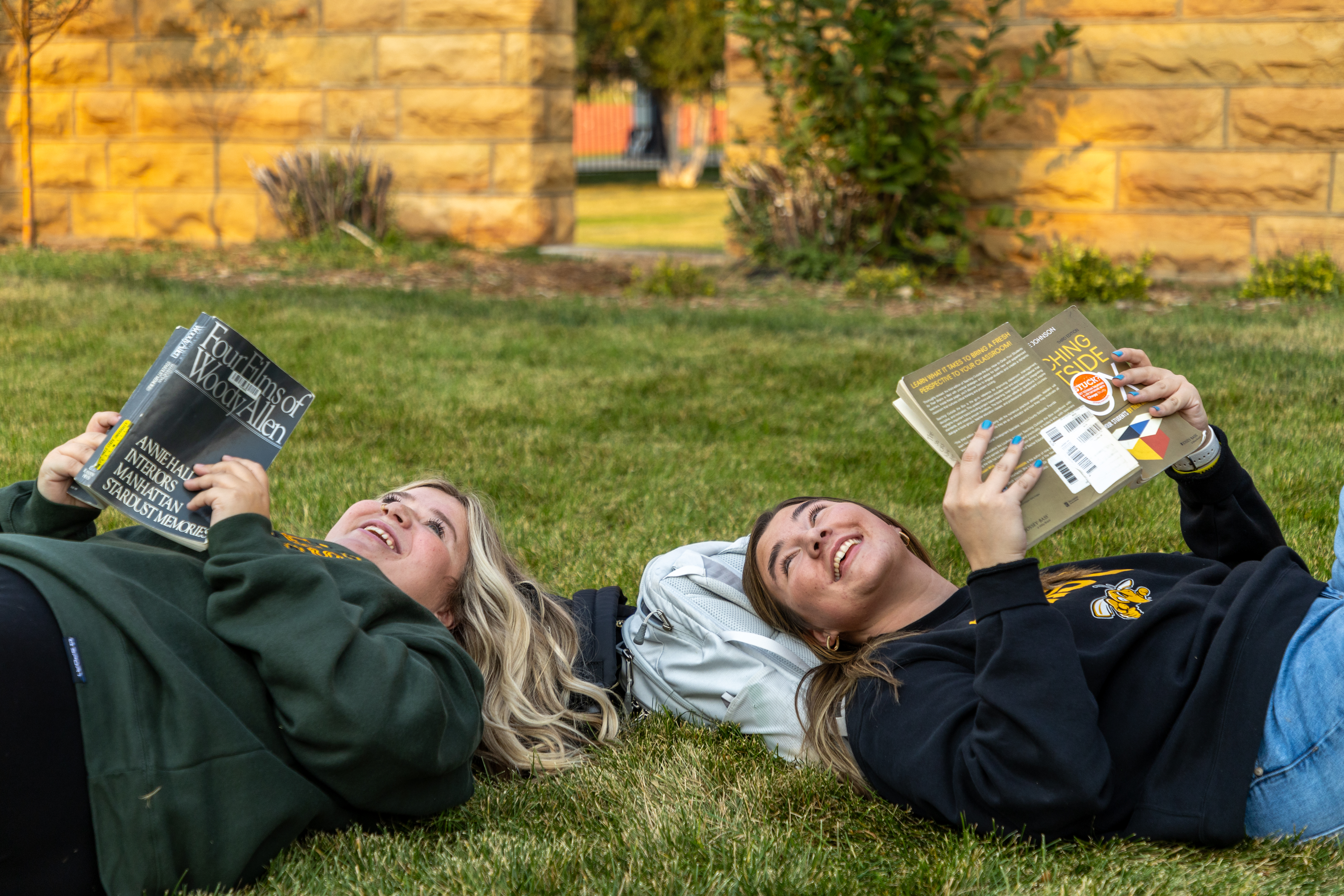 Two female BHSU students lay on the grass on campus and read.