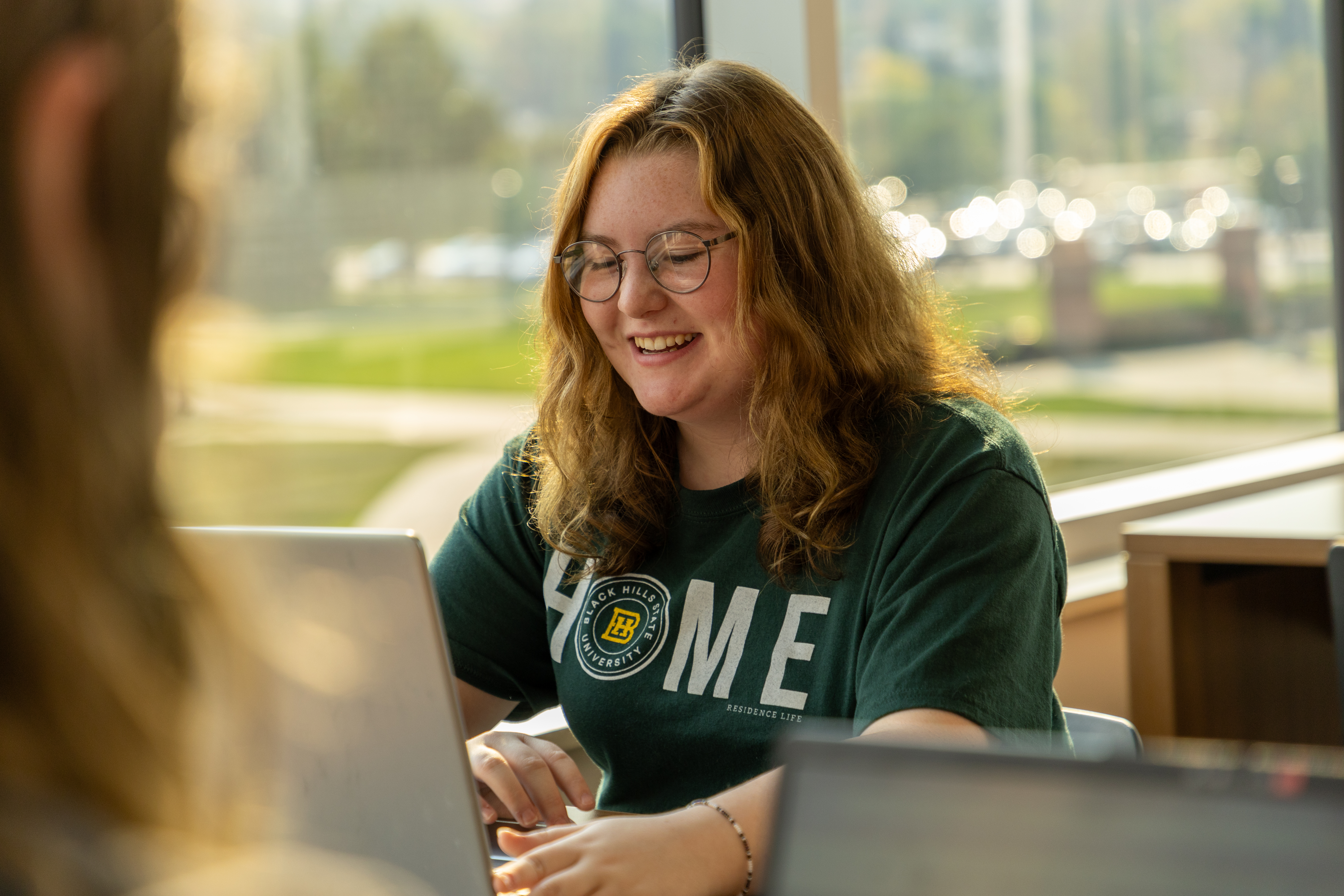 A BHSU student sits in the library in front of a window typing on a laptop.