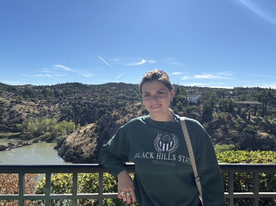A young woman with a light complexion and pulled-back hair stands smiling on a balcony overlooking a scenic landscape of rolling hills, trees, and a river. She wears a green sweatshirt that reads “BLACK HILLS STATE University.” The sky is clear and blue with wispy clouds and contrails, and green plants are visible near the railing in the foreground.