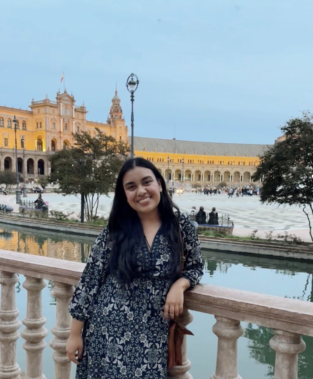 Young woman in a dark floral dress stands smiling by a decorative railing near a body of water, with a large, illuminated Spanish-style building and plaza in the background at sunset.