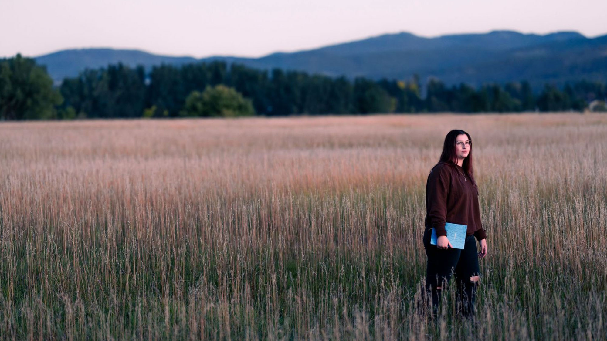 Image of Hope Schumacker in a field at sunset. There is a mountain behind her. She is holding a notebook.