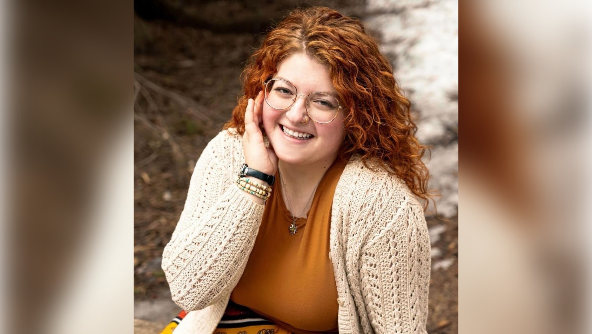  A woman with orange hair that matches her orange shirt stands outside. It appears to be autumn.  