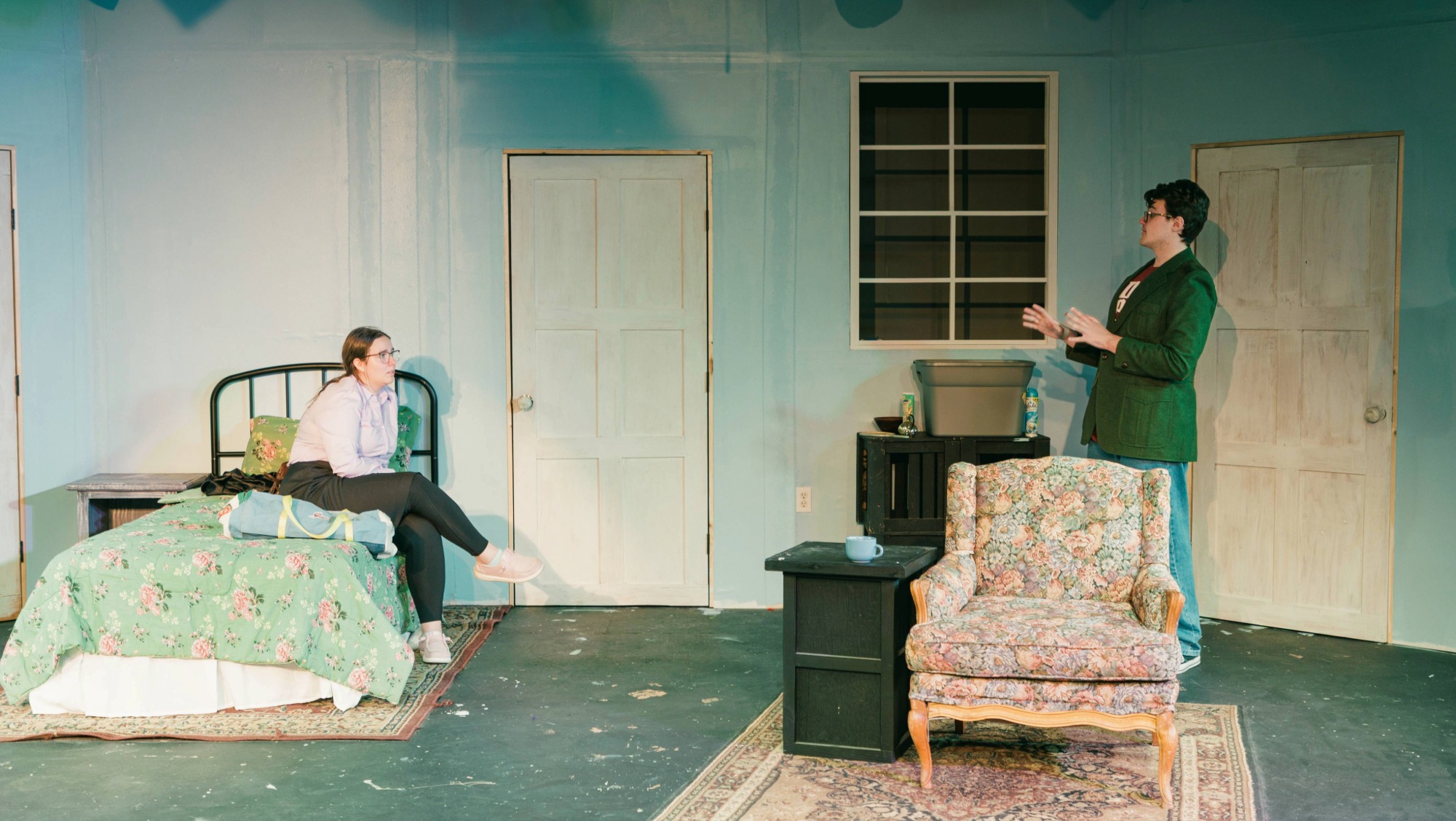 Two actors rehearse for a theatre production on a set with blue walls, a bed, and a chair