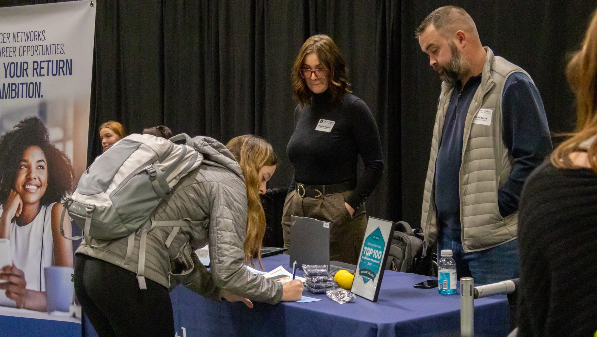 Two employers stand at their company's booth while they communicate with a student about their business