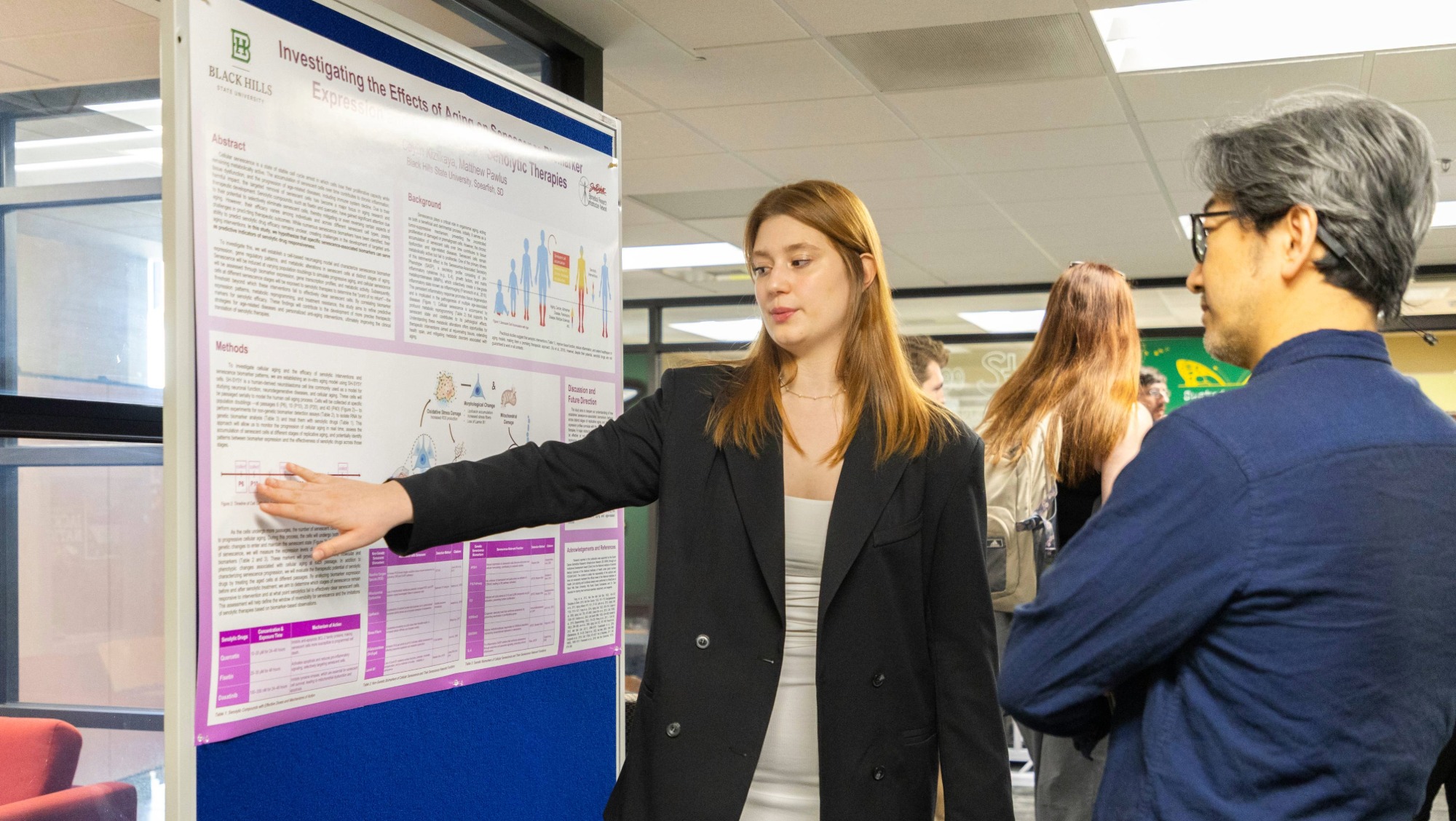 A student points to information on her poster as she presents her research to listeners
