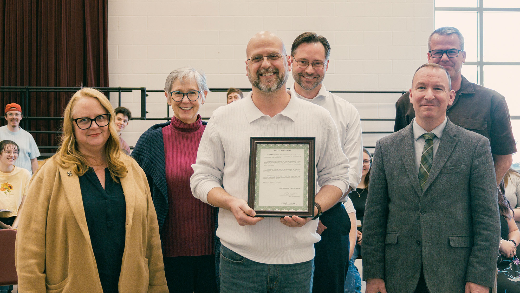 A group of six people stand together in the BHSU Choir room. Dr. Jonathan Nero in the center is holding a framed certificate and smiling. He is surrounded surrounded by others, including Dr. Pam Carriveau, provost; Dr. Amy Fuqua, dean of the College of Liberal Arts; Dr. Rich Loose, associate provost; President Steve Elliott; Quintin Owens, chair of the School of Arts. There are members of the choir in the background.