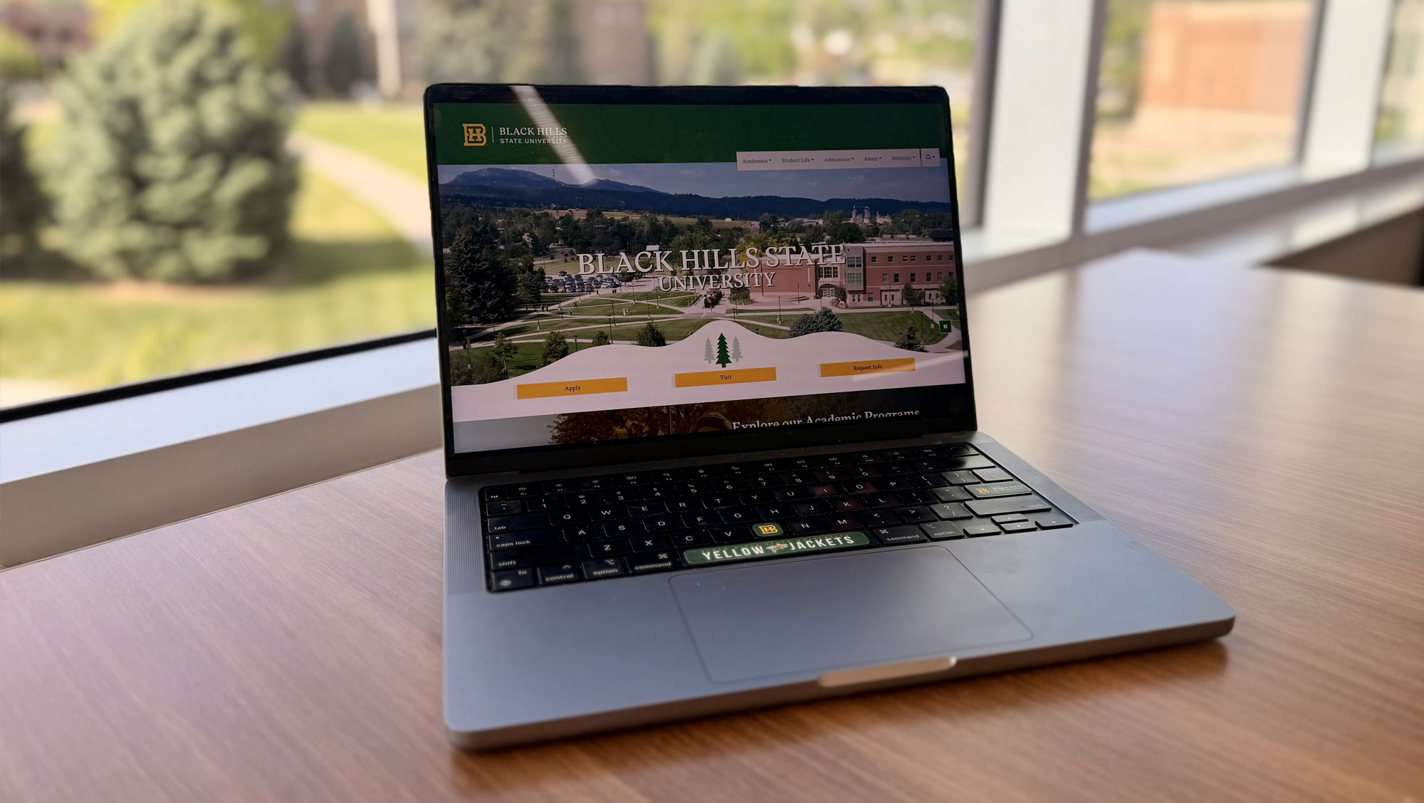 a laptop is on a desk. There is a large window behind it that shows the BHSU campus on a sunny day. The laptop has the BHSU website pulled up. It says 