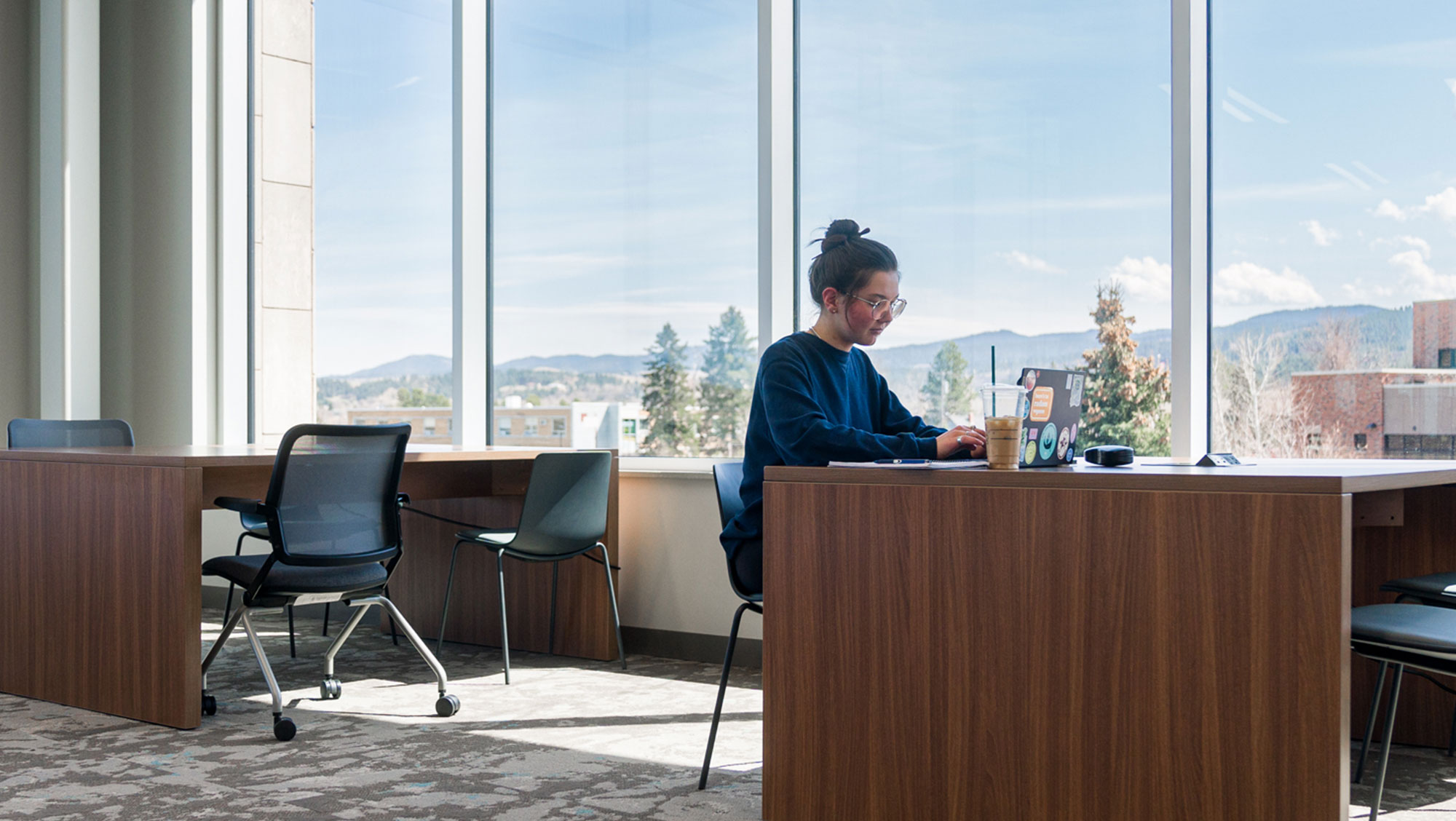 A student works on a laptop in a library with large windows. There are mountains behind her.
