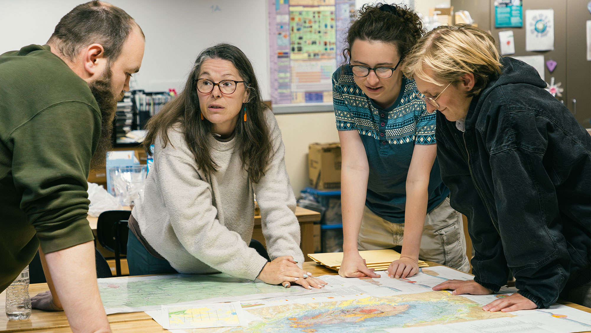 BHSU geology students and a faculty member gather around a table in a classroom, reviewing maps and documents while collaborating on a mineral exploration exercise.