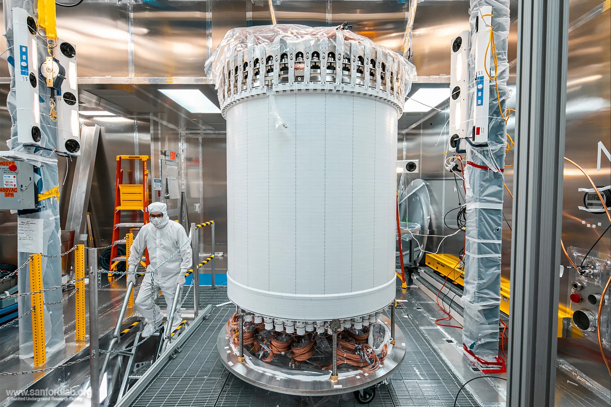 A researcher in protective gear walks down stairs near a tall, cylindrical scientific instrument with visible wiring. The walls surrounding it are reflective Teflon.