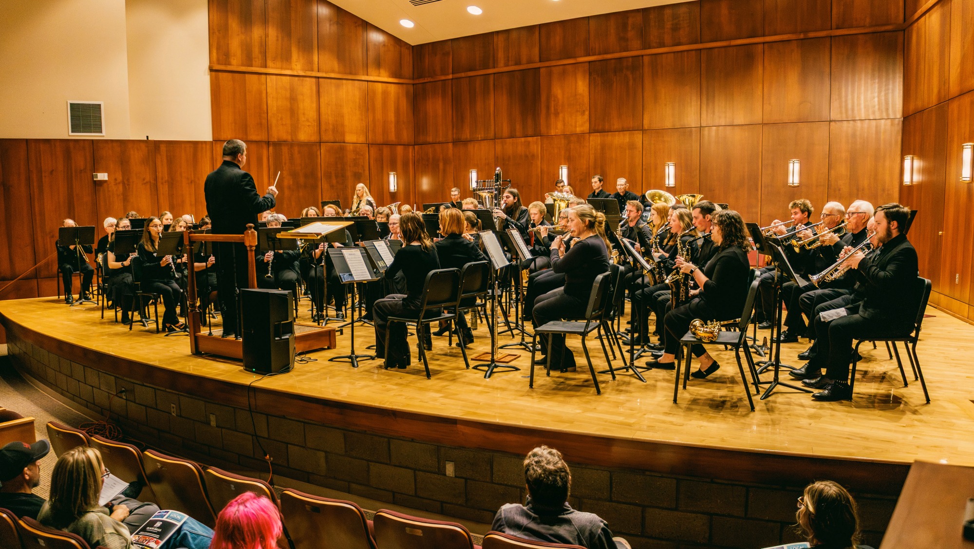 An audience watches on as the conductor stands in front of a band of people playing instruments.