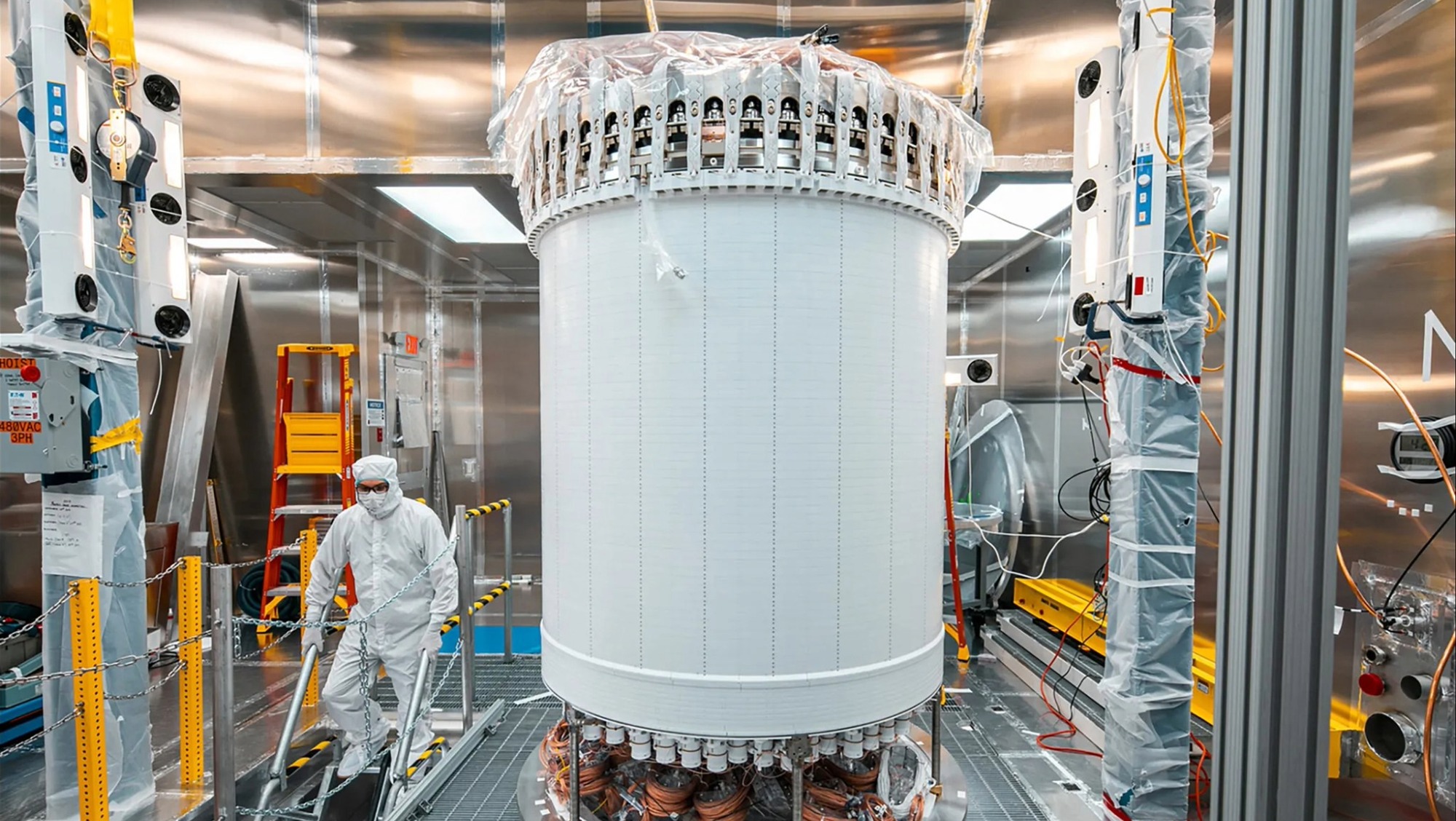 A researcher in protective gear walks down stairs near a tall, cylindrical scientific instrument with visible wiring. The walls surrounding it are reflective Teflon.