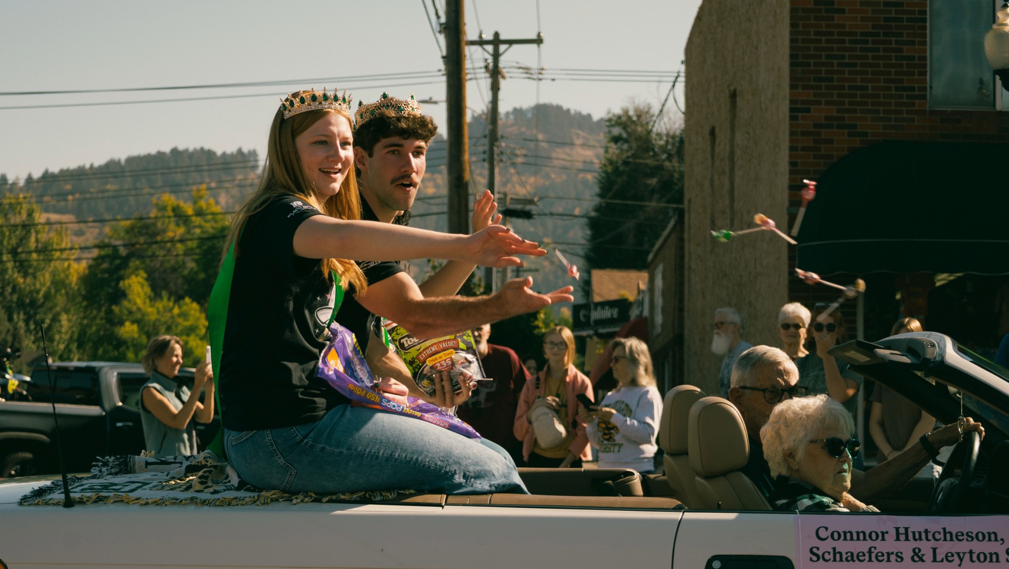 Two students wearing crowns sit in the back of a car throwing candy out during a parade 