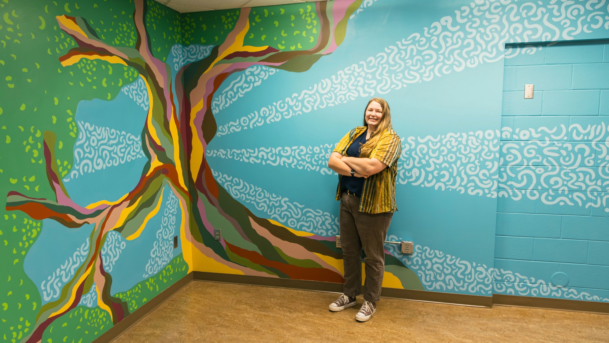 BHSU alumna Erin Peterson poses next to the large oak tree mural she designed and painted in Club Buzz in the Student Union. The mural stretches across multiple walls and features vibrant greens, blues, and earth-tone colors.