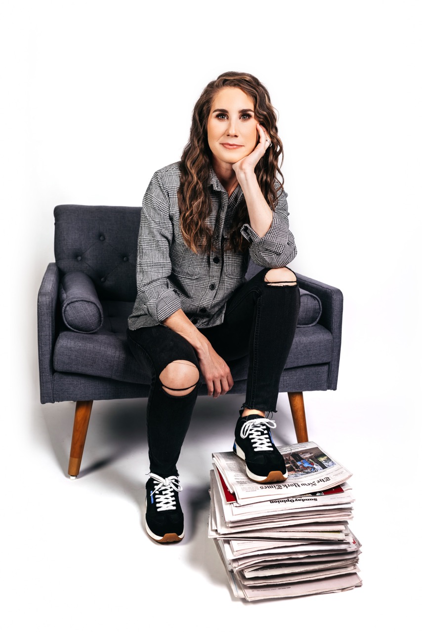 Person sitting in a gray armchair, resting their head on one hand, wearing a plaid shirt, black ripped jeans, and black sneakers, with one foot placed on a stack of newspapers against a white background.