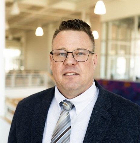 Person wearing glasses, a dark blazer, white shirt, and striped tie, standing indoors in a bright modern building with blurred lights in the background.