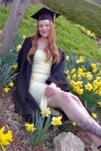 Graduate Olivia Schaefers poses in her cap and gown surrounded by yellow flowers