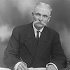 Black and white image of William J. Collins signing a document while sitting at a desk.