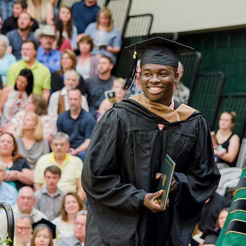 Picture of BHSU graduate accepting his diploma; large audience in the background sitting in the bleachers of the Young Center Gym.